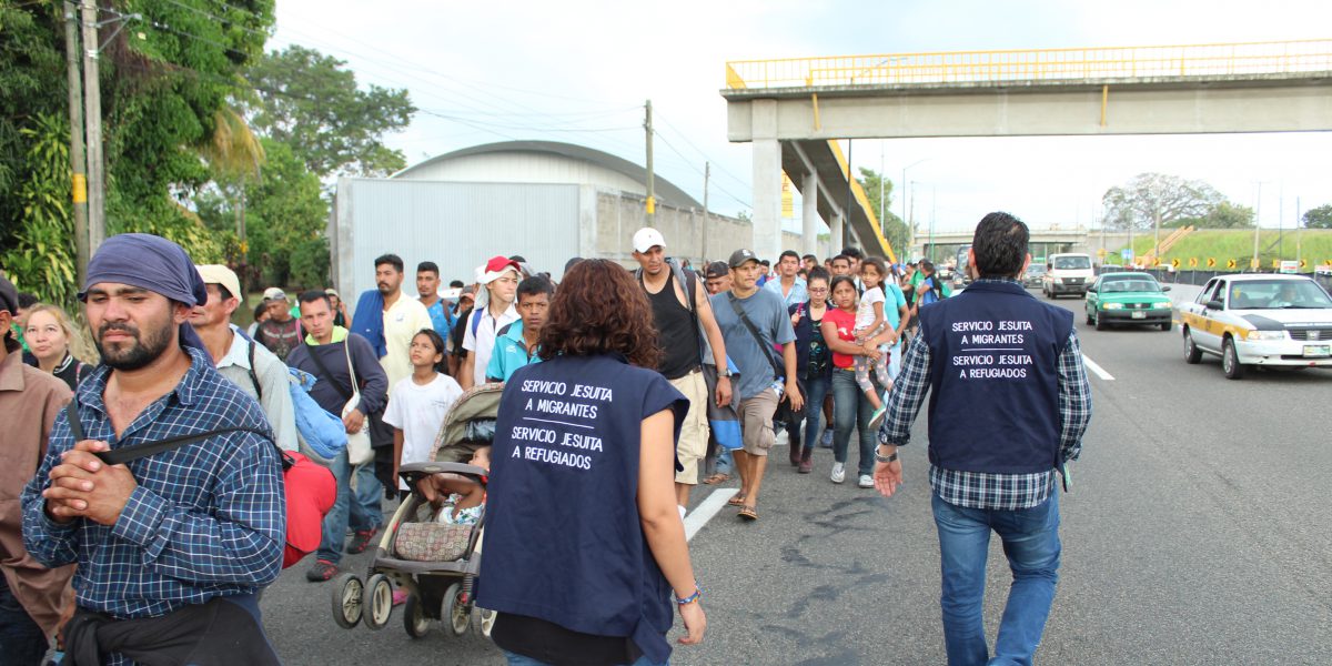 A variety of people walking on the left side of the street. Some are carrying luggage, and others are wearing blue JRS vests, assisting those in transit.