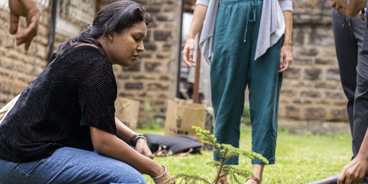 A woman bent over planting a tree while people stand around with shovels.