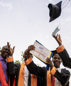 Four young men in graduation caps and gowns, one holding a diploma and one tossing his graduation cap in the air. All are smiling.