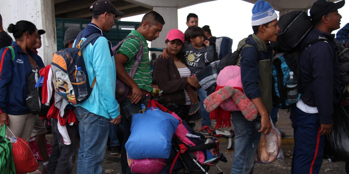 A group of women, men, and children all carrying backpacks and other luggage standing and waiting in line to get transportation.