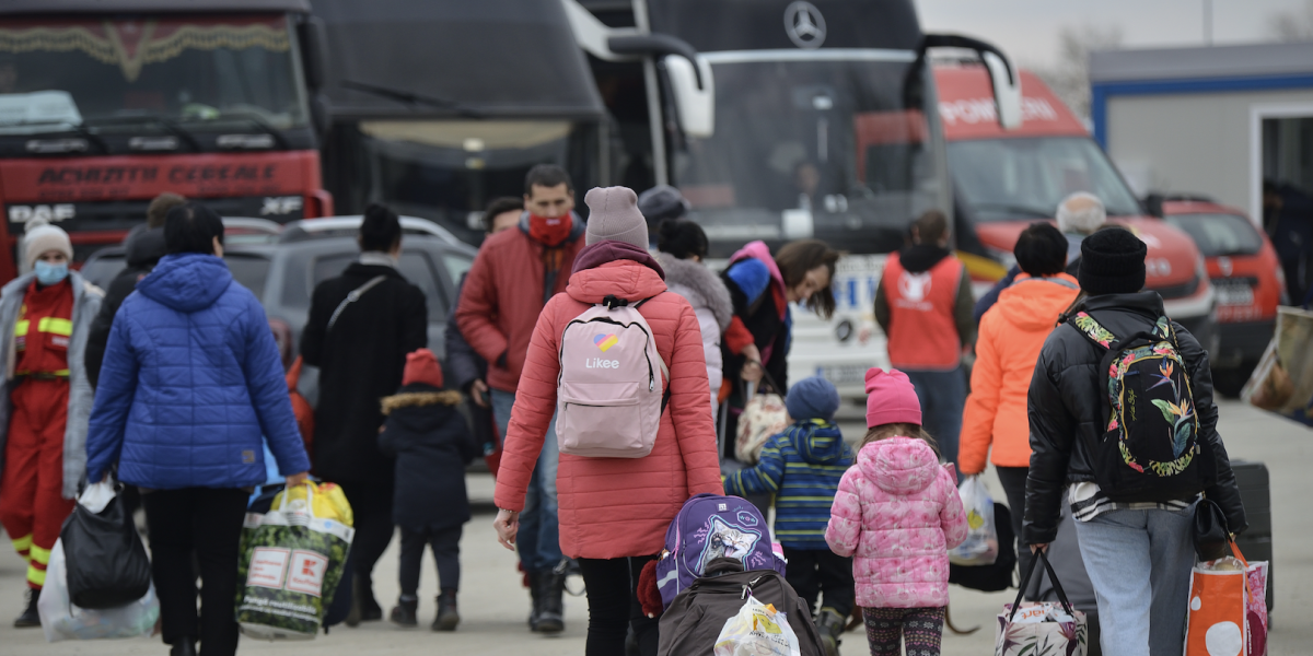 A group of people walking towards busses and cars while carrying luggage.