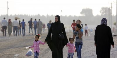 A woman in a burqa walking outside holding the hands of three children. Two of the children are younger and wearing pink tops, one is an adolescent wearing a light blue top and jeans with a baby sitting on her shoulders. There is smog in the background.