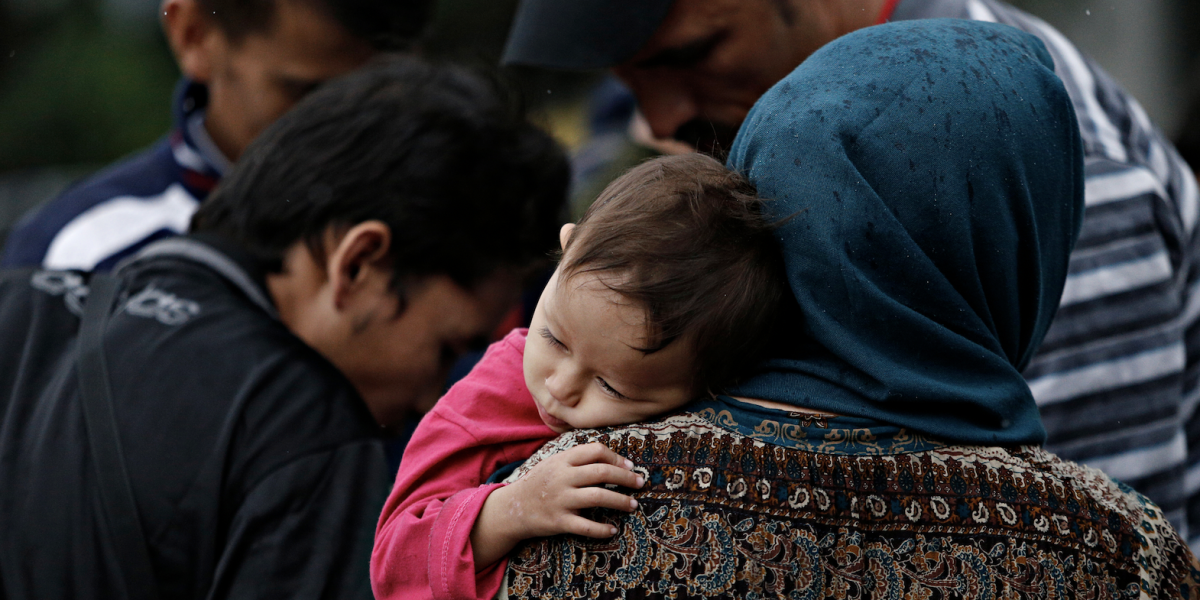 A young child laying on a mother's shoulder with her back turned towards the camera. There are various people in the background as well.