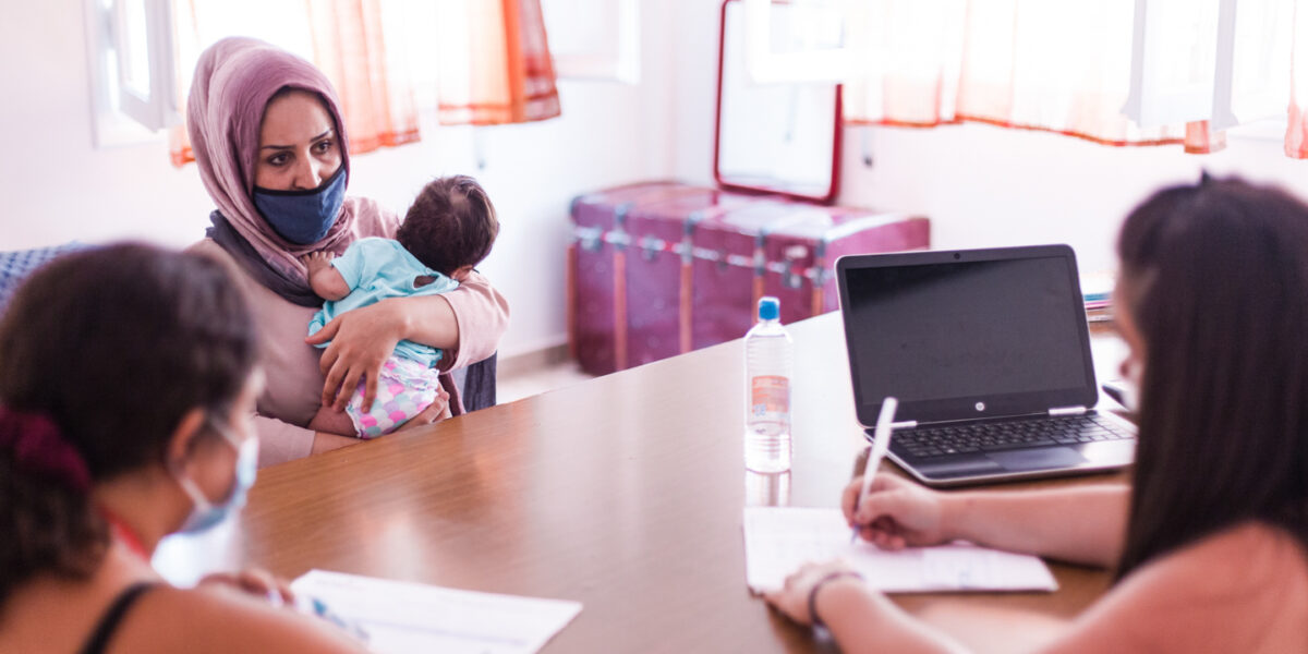 A woman holding a small infant is speaking across a brown desk to two other women who appear to be taking notes. The woman is receiving some type of medical advice.
