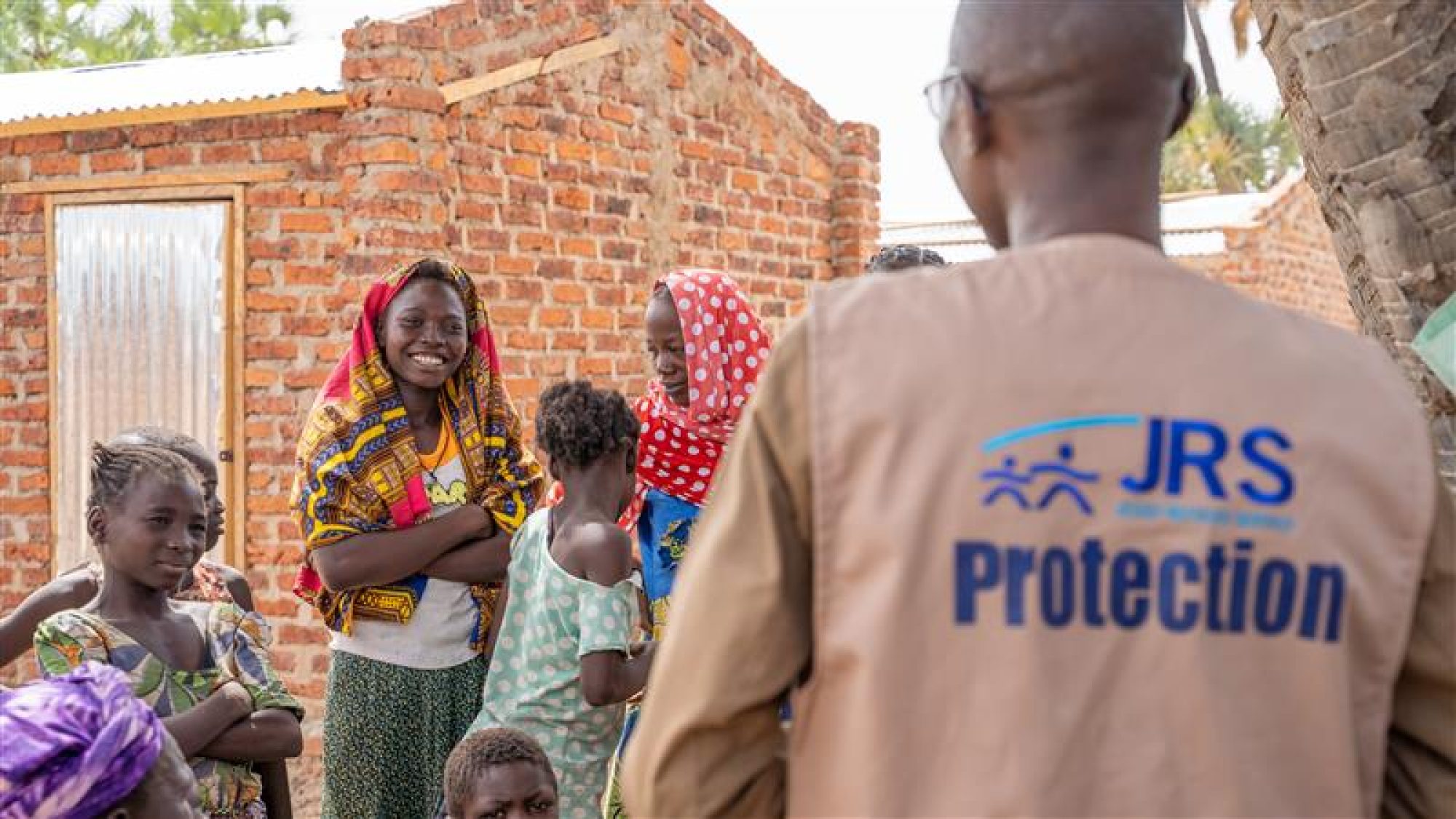 World Humanitarian Day: Why humanitarian aid is vital to communities everywhere. A JRS staff member who is wearing a JRS vest that says 'Protection,' with his back facing the camera while looking at a group of women and girls who are smiling at him.