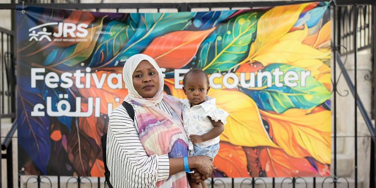 JRS Jordan Festival of Encounter. A mother. stands with her baby in front of a JRS Jordan Festival of Encounter sign. This festival is on its third year in a row and celebrates the diversity of the refugee community in Amman, Jordan.