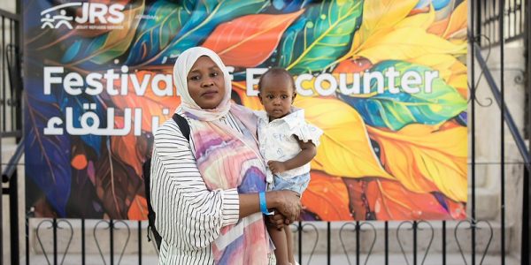 JRS Jordan Festival of Encounter. A mother. stands with her baby in front of a JRS Jordan Festival of Encounter sign. This festival is on its third year in a row and celebrates the diversity of the refugee community in Amman, Jordan.