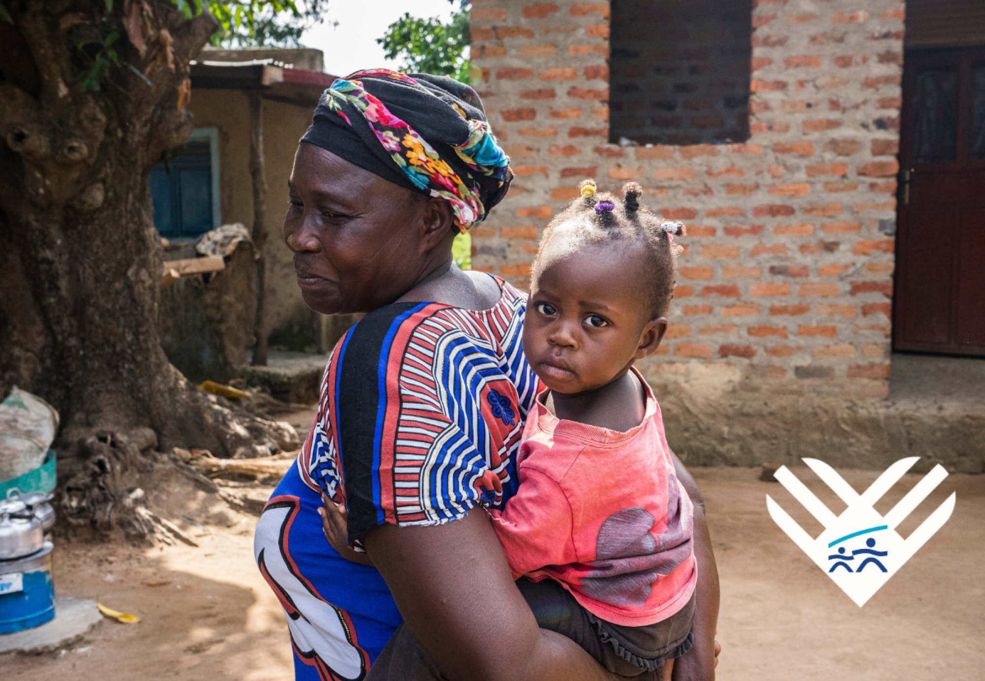 JRS Uganda in Adjumani and Moio. A mother holding her child on her back in front of a stone shelter. Support JRS this Giving Tuesday—give vital food support to malnourished children and pregnant mothers.