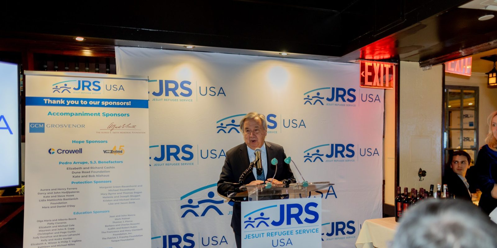 United Nations Secretary-General António Guterres Honored at JRS/USA 45th Anniversary Dinner. A man standing at a podium speaking to a small, intimate crowd. He is standing in front of a banner with the JRS/USA logo on it.