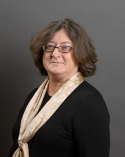 A woman with chin-length brown hair and glasses, wearing a black top and a cream-colored scarf, sitting in front of a grey background. | Ignatian Migration Dialogue, State and Local Immigration Measures