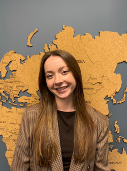Chloe Gunther smiling at the camera, wearing a tan plaid blazer, standing in front of a cork world map.
