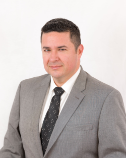 A man with dark hair wearing a grey suit with a black tie, sitting in front of a white background staring at the camera. Ignatian Migration Dialogue, State and Local Immigration Meausres