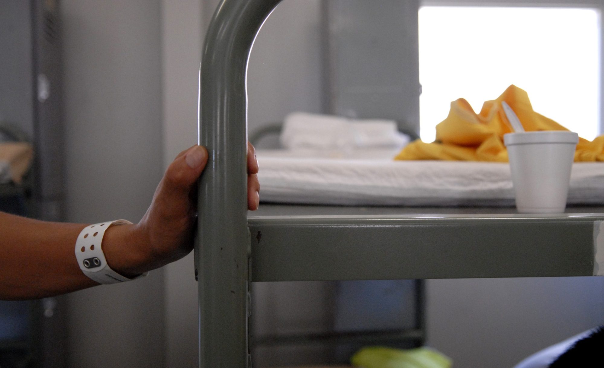 A minimal cot with a hand holding it. The hand has a tag bracelet on the wrist. Contact the White House to Change Course on Harmful Deportation Practices
