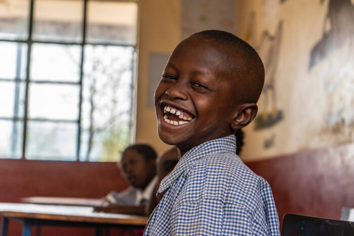 A child sitting in a classroom looking back at the camera with a big smile and hope in his eyes. There is a window with light shining through behind him.