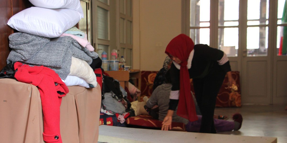 A displaced immigrant kneeling down in a church with several piles of clothes around. There is a thin mattress on the floor.