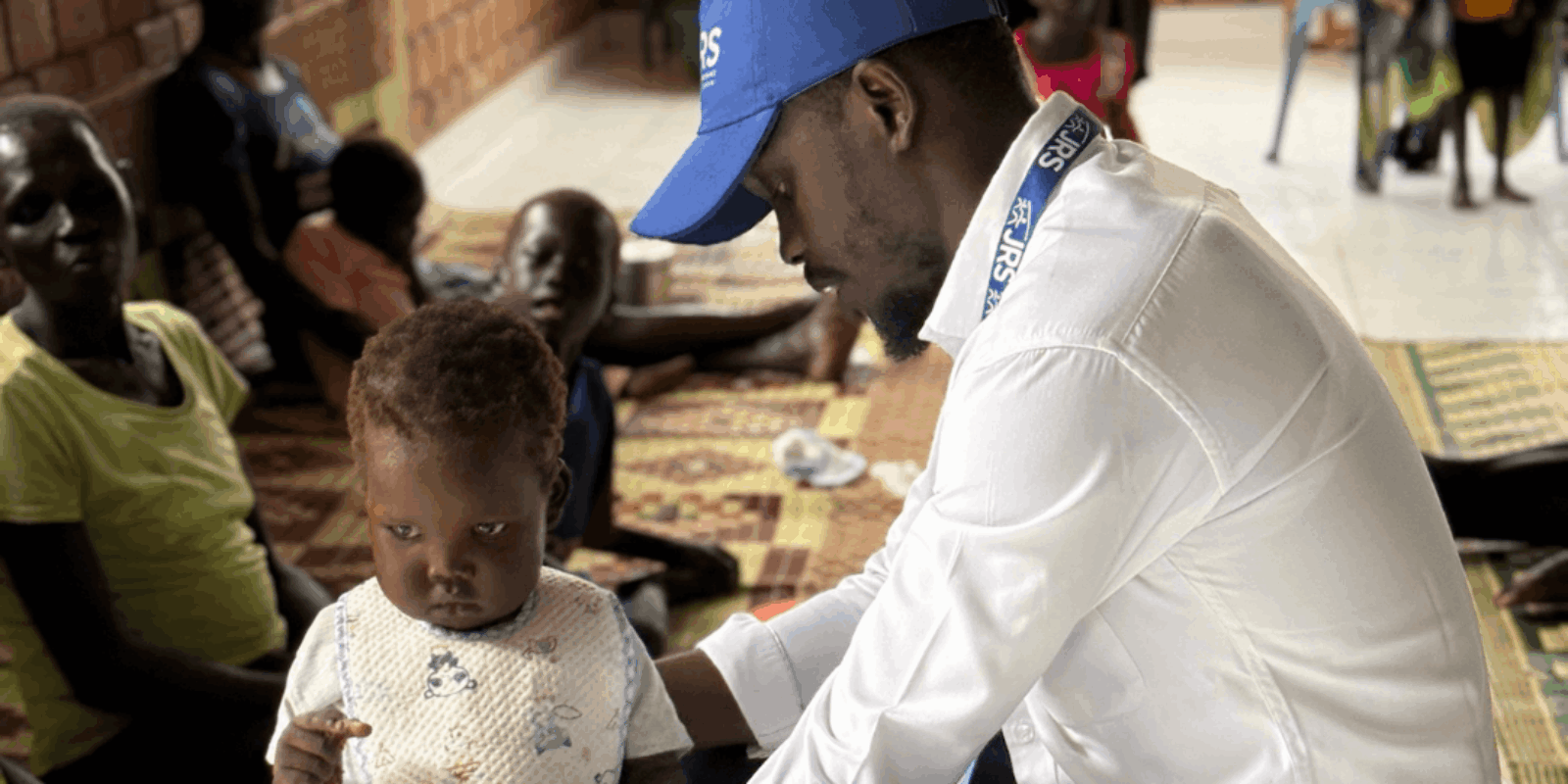 A man is crouched down supporting and observing a small child who is walking. The man is wearing a blue JRS hat, a white shirt, and black pants. The child is wearing a white top and white pants. They are in a room with other people sitting against a wall.