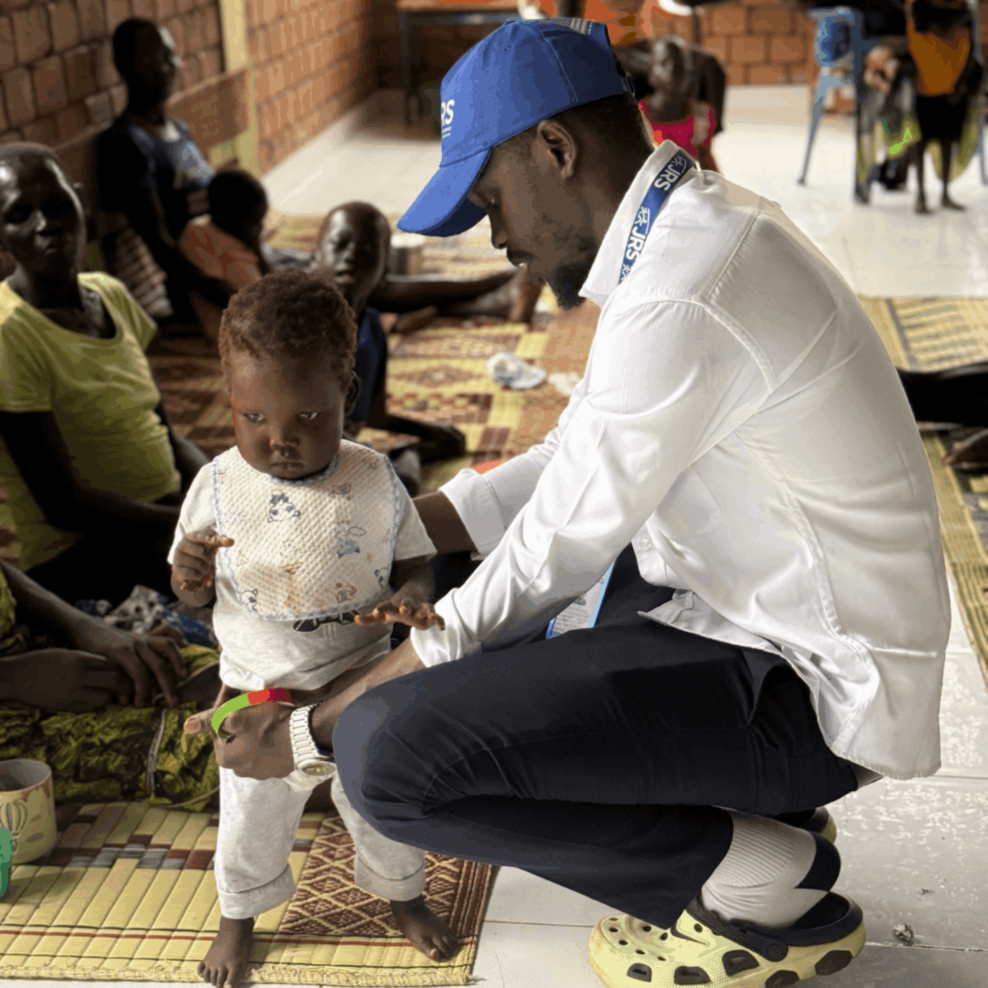 A man is crouched down supporting and observing a small child who is walking. The man is wearing a blue JRS hat, a white shirt, and black pants. The child is wearing a white top and white pants. They are in a room with other people sitting against a wall.