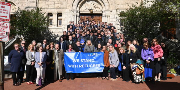 A group of people standing together on church steps holding a blue banner that says 'We Stand with Refugees' with the JRS/USA logo in the top right.