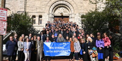A group of people standing together on church steps holding a blue banner that says 'We Stand with Refugees' with the JRS/USA logo in the top right.