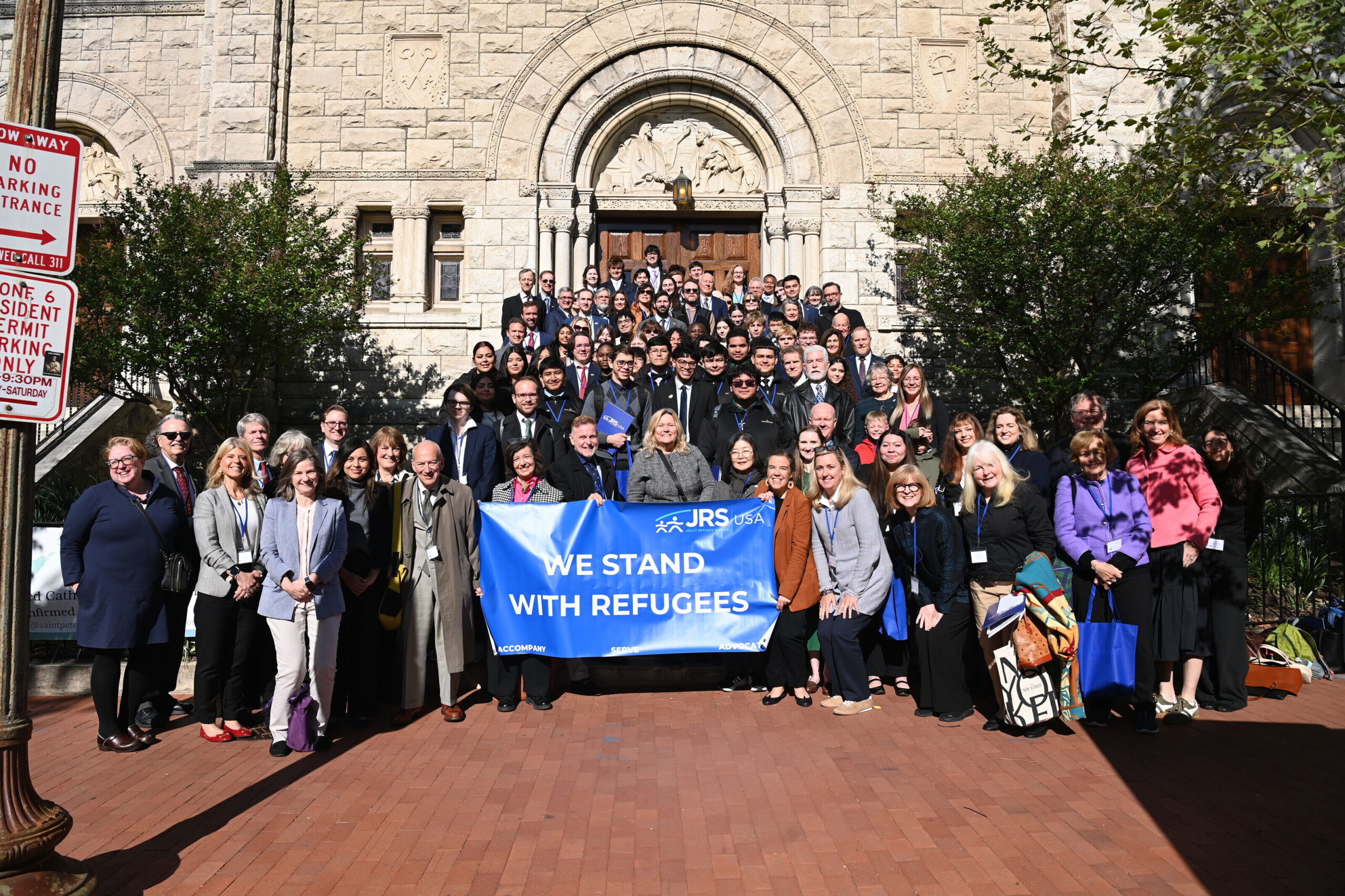 A group of people standing together on church steps holding a blue banner that says 'We Stand with Refugees' with the JRS/USA logo in the top right.