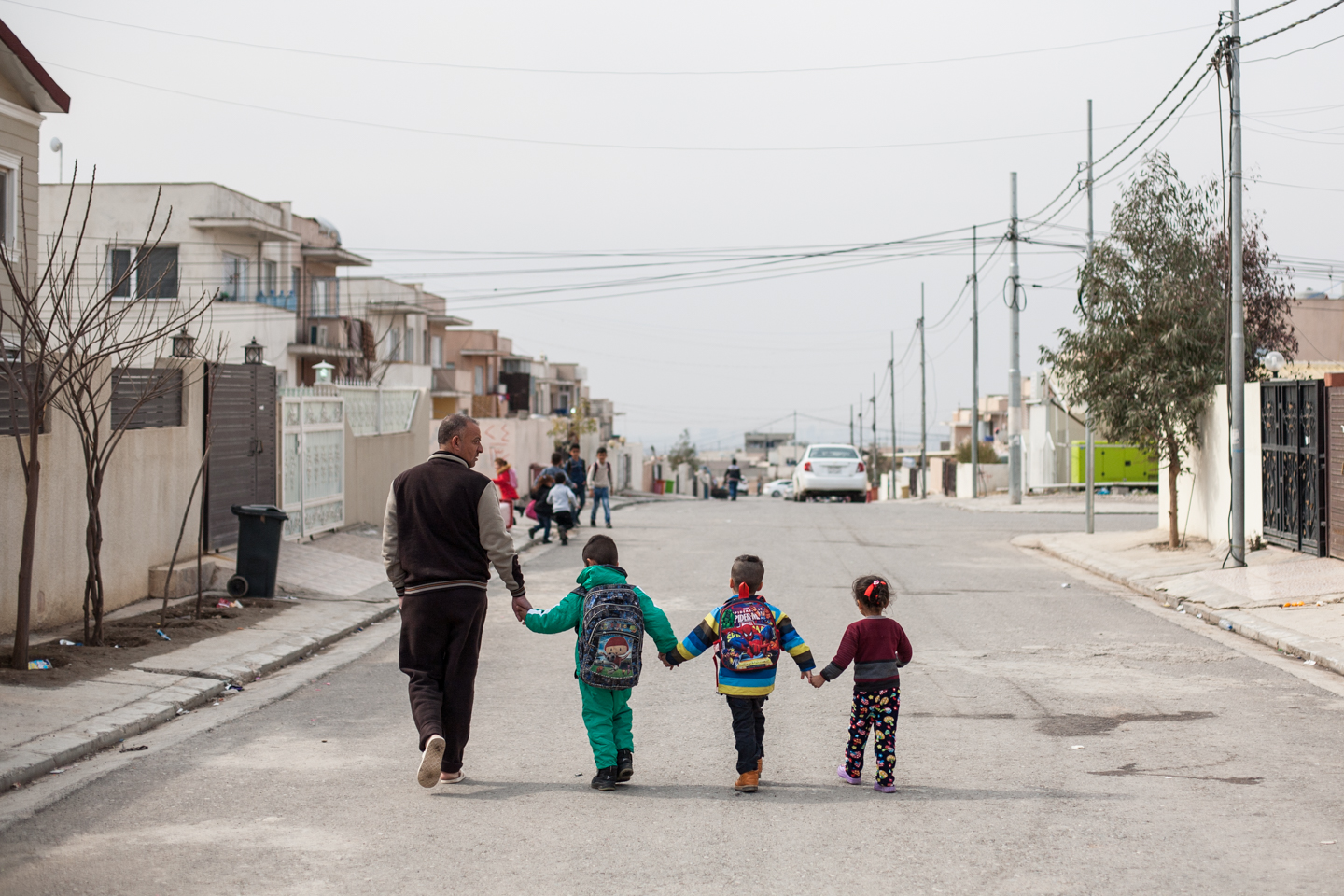 A family of four walking down the middle of an empty street while holding hands. There appears to be one adult and three children. They all have their backs turned to the camera facing a grey sky.