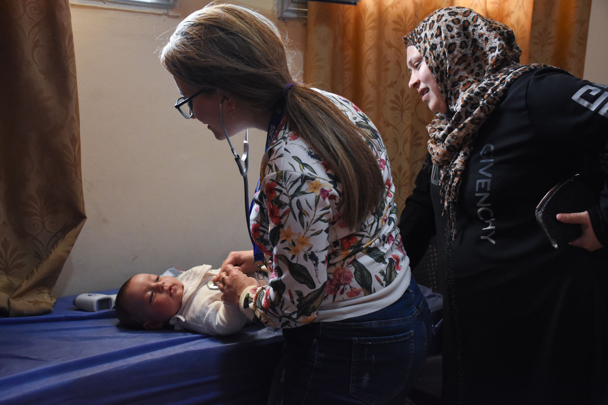 Doctor listening to an infant with a stethoscope. The infant looks content and the mother is standing behind the doctor watching. Support the MERCY Act today.