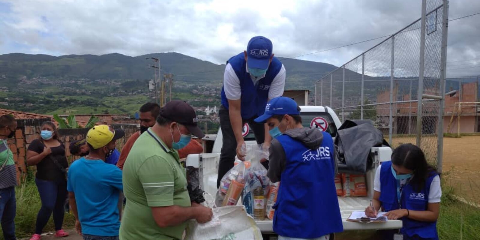 JRS humanitarian aid workers supporting refugees and other forcibly displaced people in Venezuela. There are people wearing blue vests with JRS on them, handing people different food items from a truck.