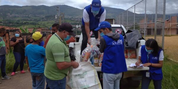 JRS humanitarian aid workers supporting refugees and other forcibly displaced people in Venezuela. There are people wearing blue vests with JRS on them, handing people different food items from a truck.
