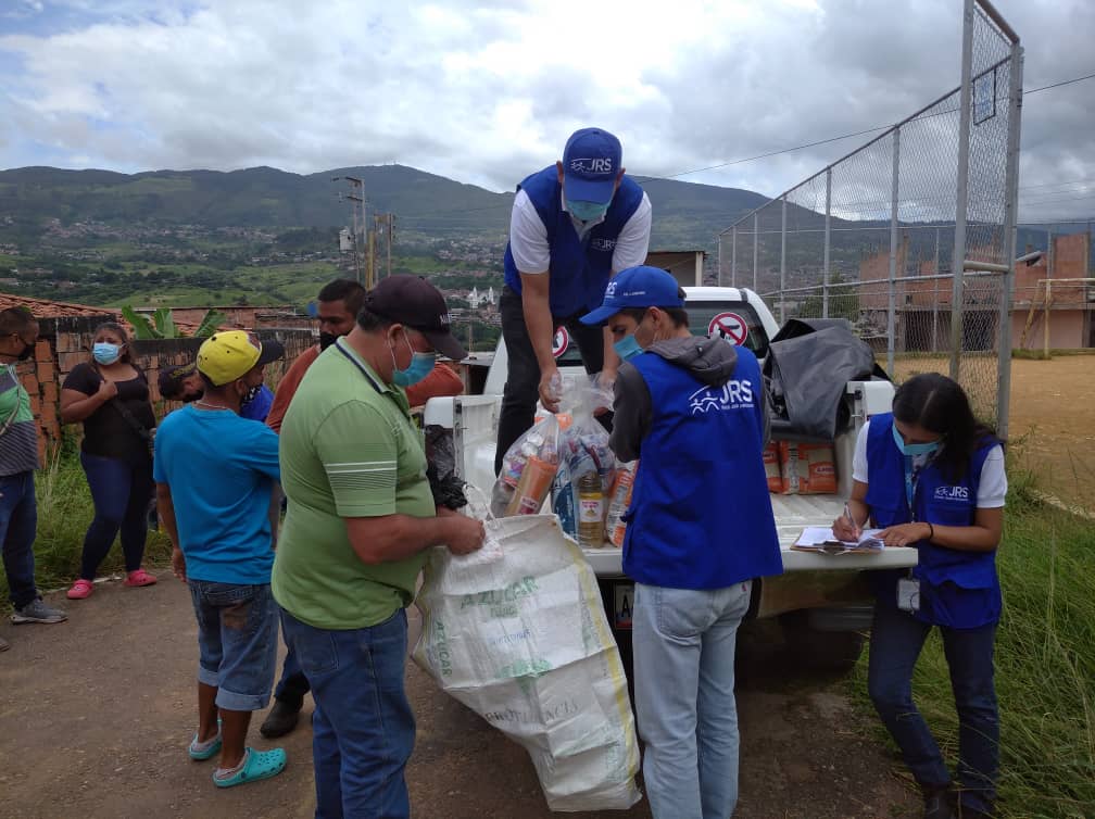 JRS humanitarian aid workers supporting refugees and other forcibly displaced people in Venezuela. There are people wearing blue vests with JRS on them, handing people different food items from a truck.