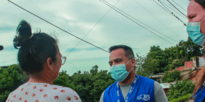 A man wearing a mask and a blue vest with 'JRS' on the top right. He is shaking hands with a woman who has her back turned towards the camera.